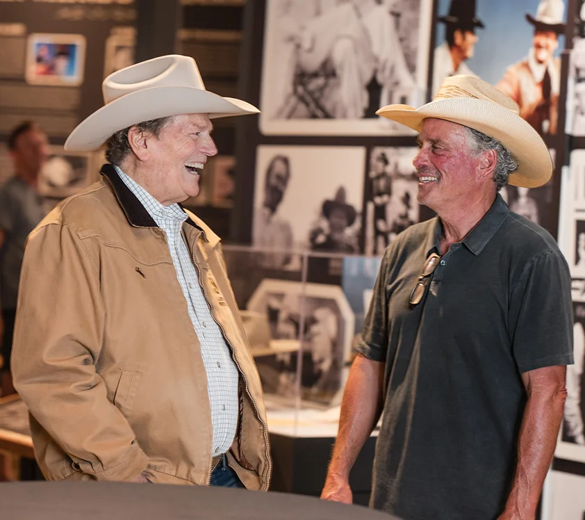 man talking to other man laughing in front of a john wayne exhibit